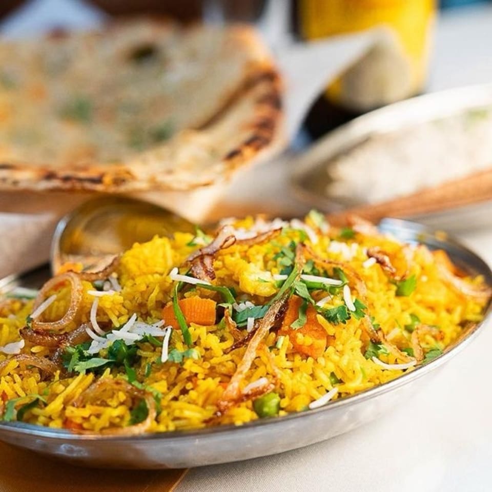 A platter of saffron-colored biryani rice garnished with herbs, fried onions, vegetables, and flatbread in the background.