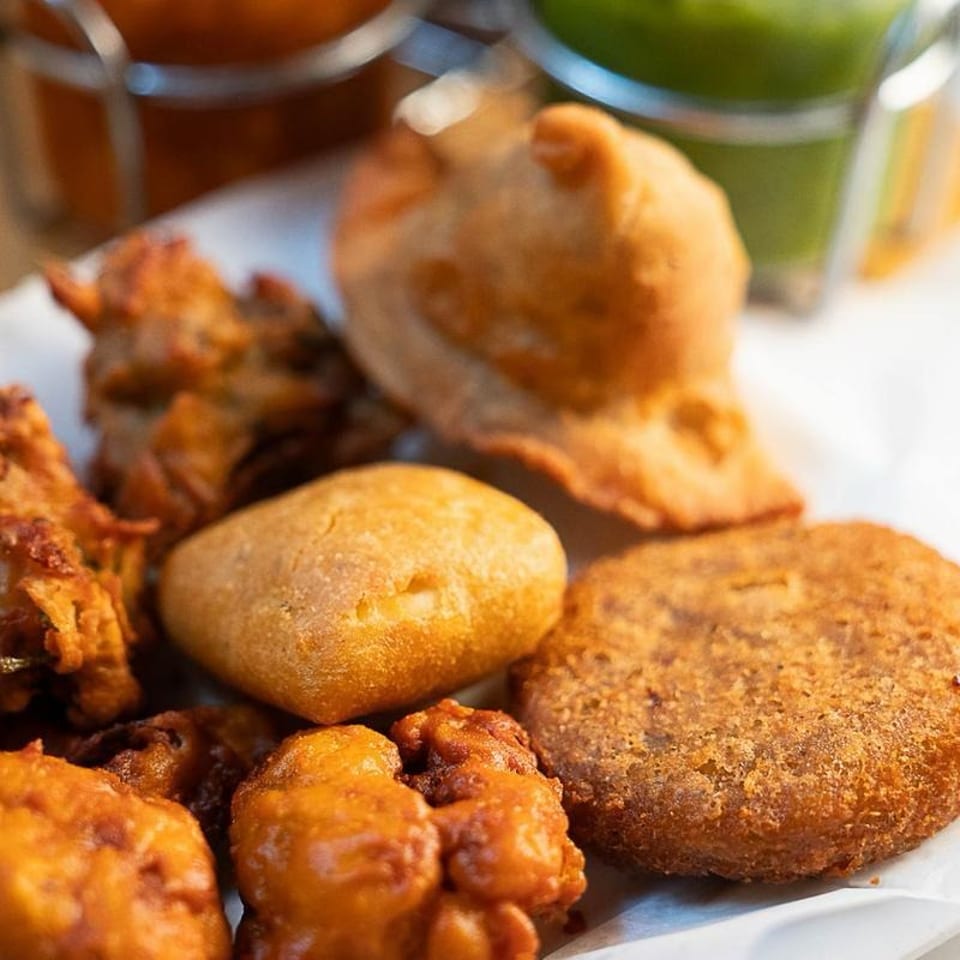 Assortment of golden fried pastries and savory snacks served on a plate, with colorful drinks blurred in the background.