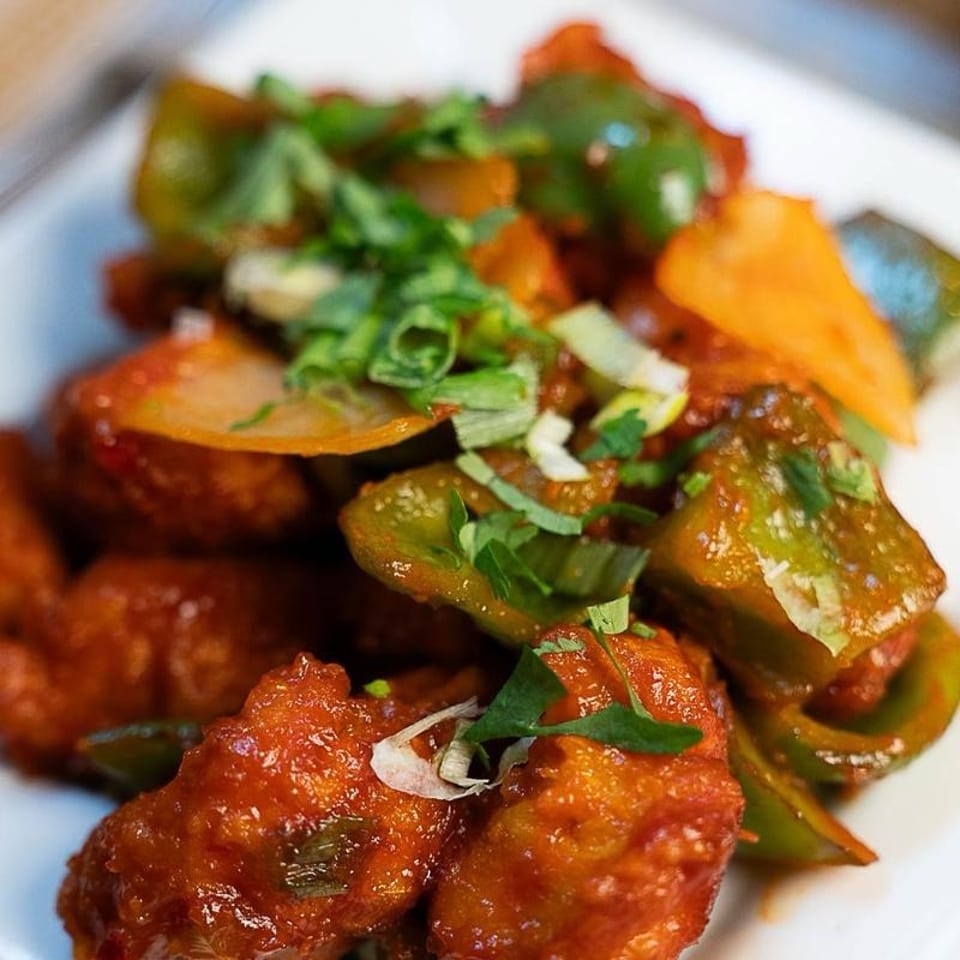 Close-up of crispy, red-glazed fried pieces topped with sautéed peppers, onions, and fresh herbs on a white plate.