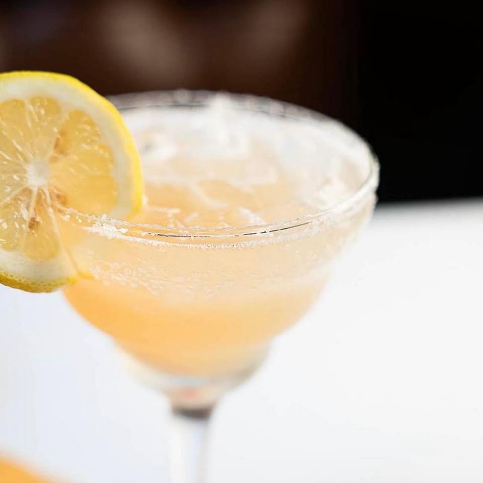 Pale orange cocktail in a stemmed glass with a salted rim and lemon slice garnish.
