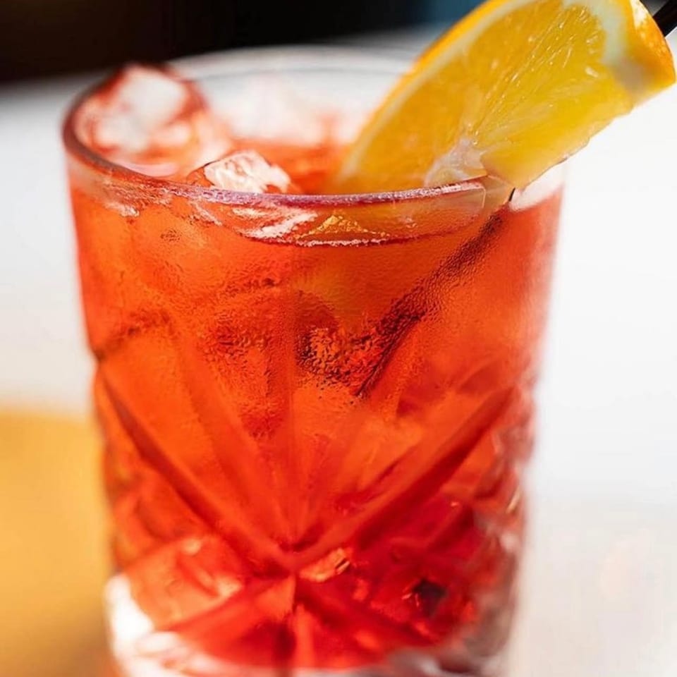 Refreshing red-orange cocktail served over ice in a faceted glass, garnished with a fresh orange wedge.