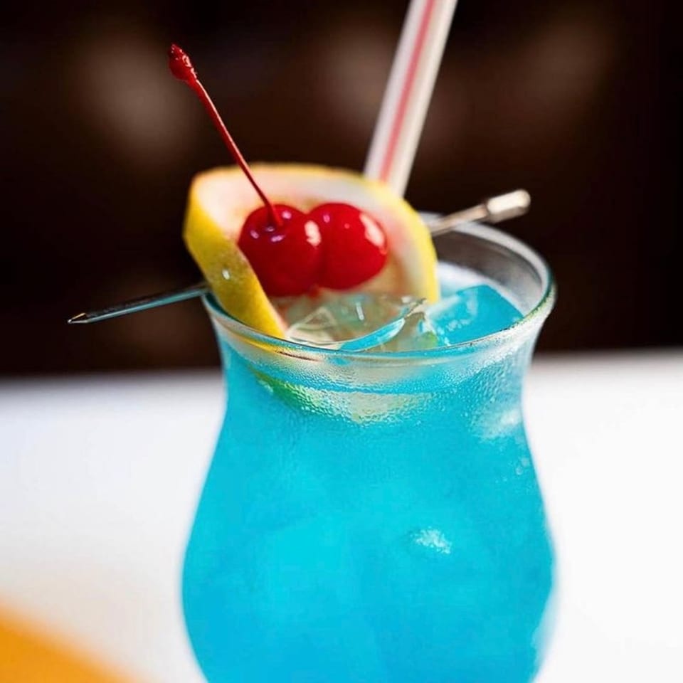 Bright blue cocktail in a stemless glass, garnished with a lemon wedge and red cherries, with a straw and pick.